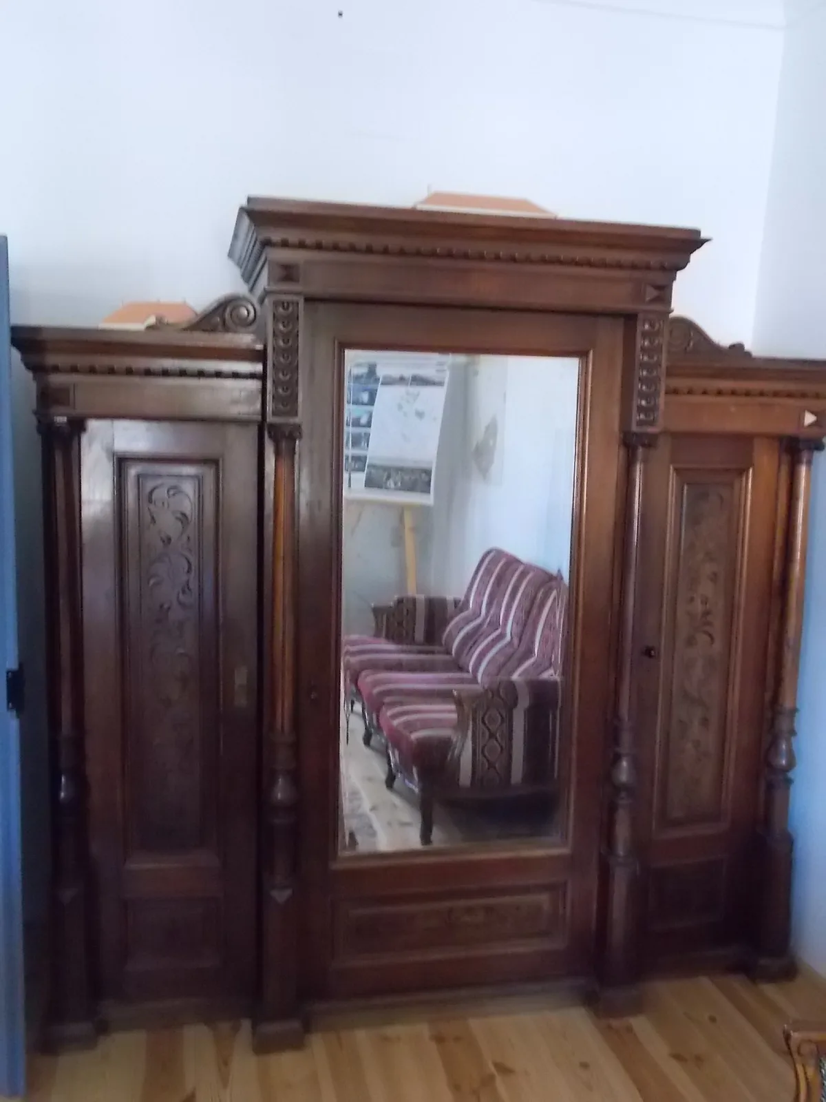Een houten kast met een grote spiegel in het middenpaneel, waarin een kamer met gestreepte fauteuils en een deur weerkaatst wordt.