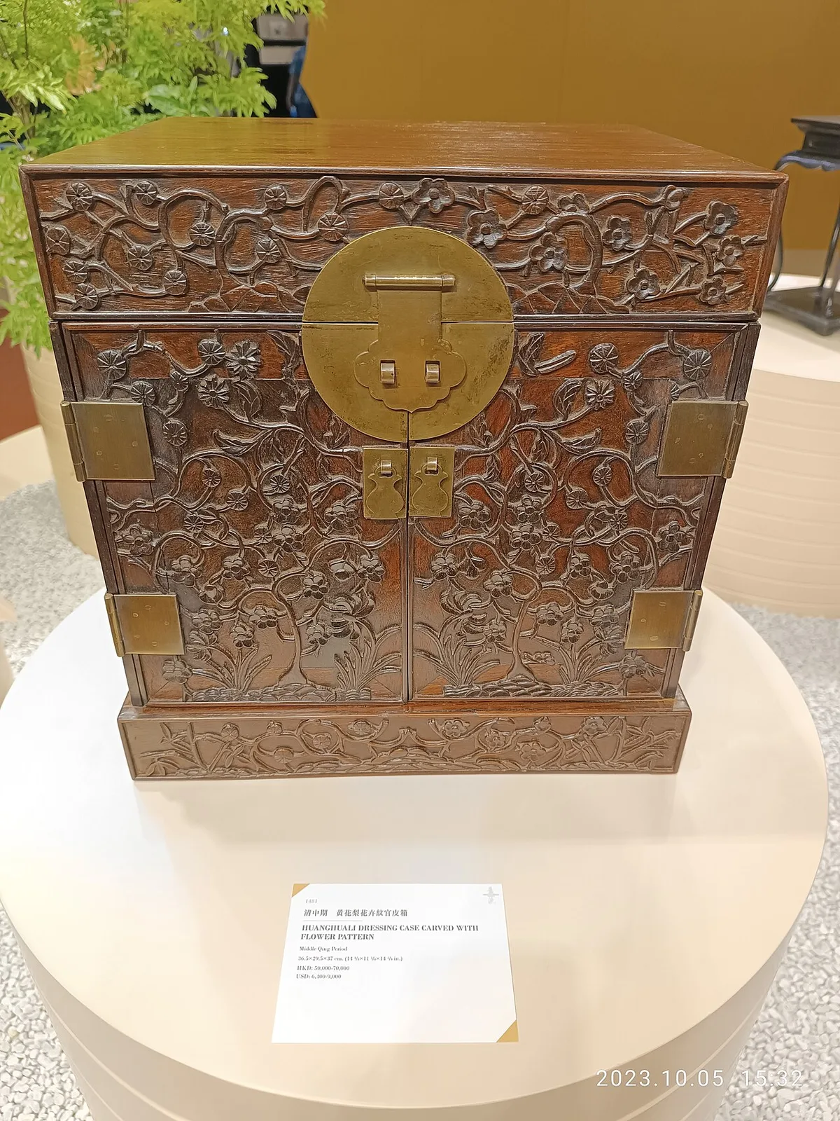 Een antieke houten kast met fijn houtsnijwerk en messing beslag, opgesteld op een witte sokkel in een tentoonstellingsruimte.